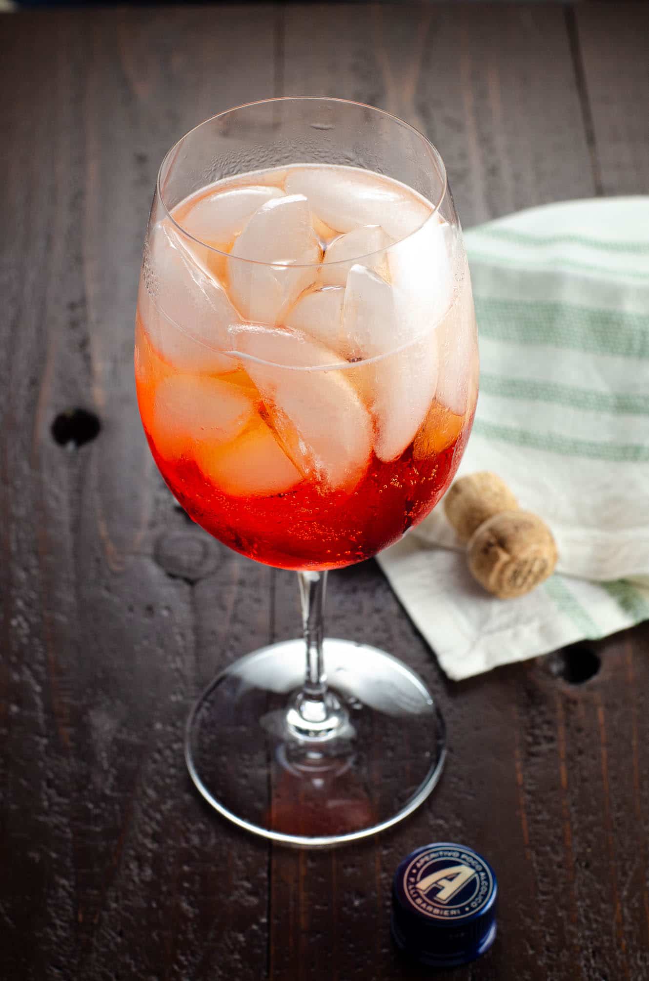 an aperol spritz (3 2 1 recipe) in a large wine glass on a wooden table with a cork and a napkin