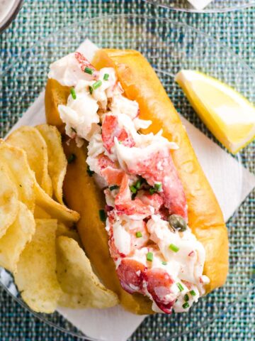 the best lobster roll recipe on a plate with potato chips, a lemon wedge, and a beer