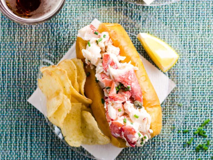 the best lobster roll recipe on a plate with potato chips, a lemon wedge, and a beer