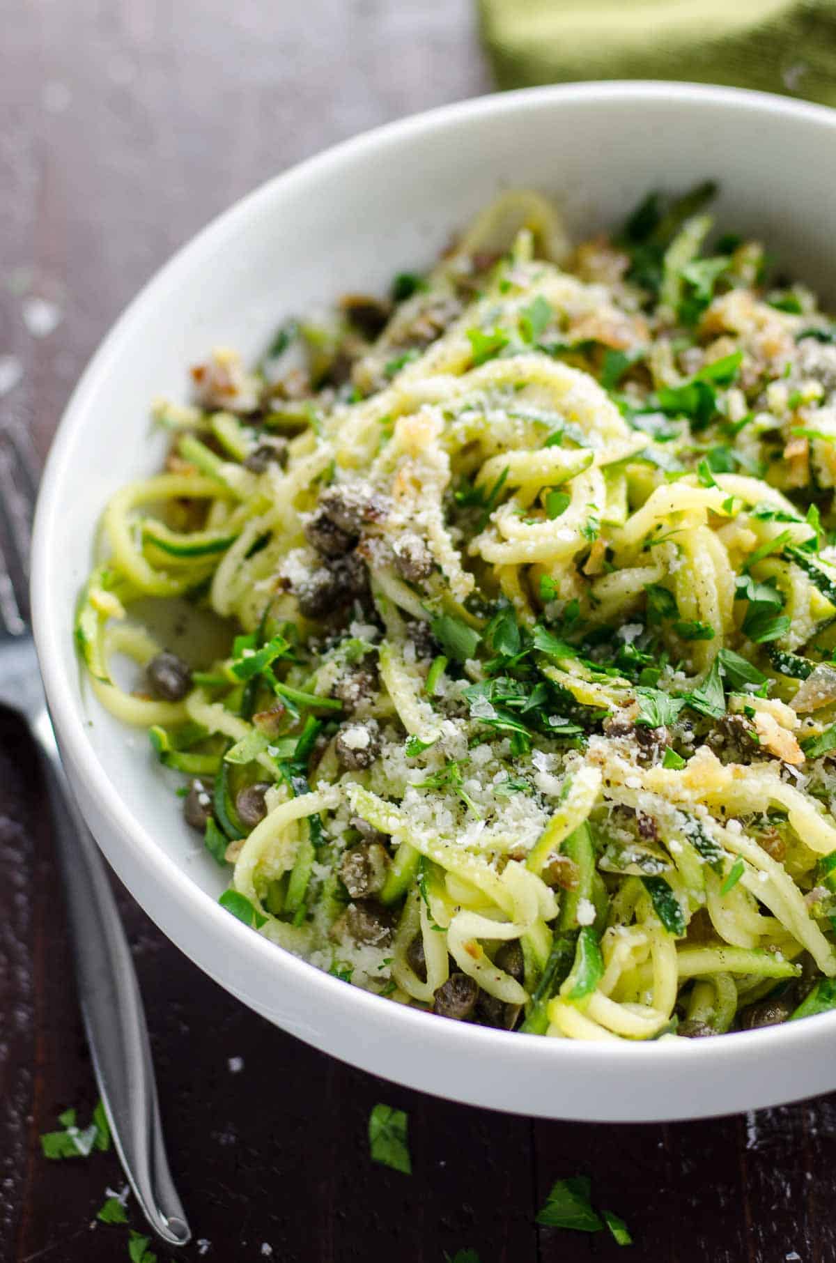 zoodles recipe with lemon caper butter in a white bowl with a fork and green napkin