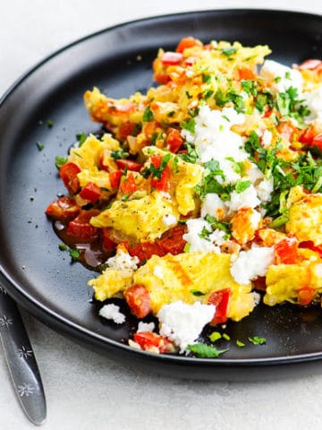 scrambled eggs cooked with onion and red bell pepper, sprinkled with cotija cheese, cilantro, and hot sauce, on a black plate on a white background