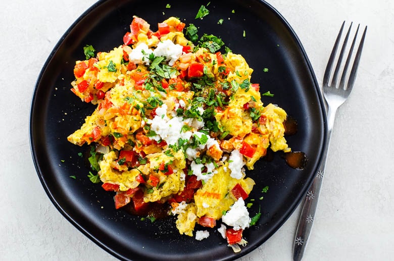 overhead shot of scrambled eggs cooked with onion and red bell pepper, sprinkled with cotija cheese, cilantro, and hot sauce, on a black plate on a white background