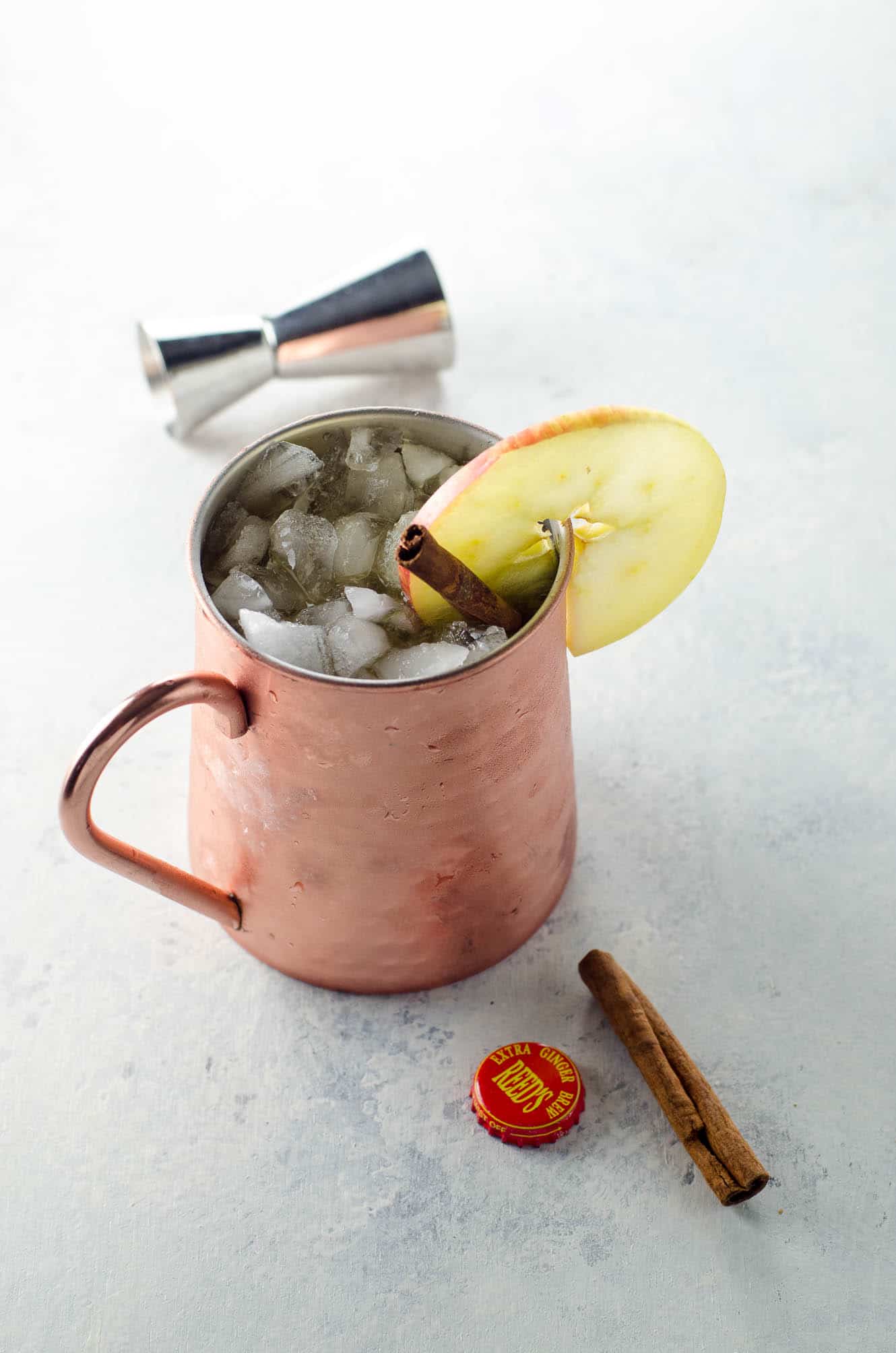 an apple cider moscow mule in a copper mug, garnished with an apple slice and a cinnamon stick, with a ginger beer bottle cap and a jigger on the table