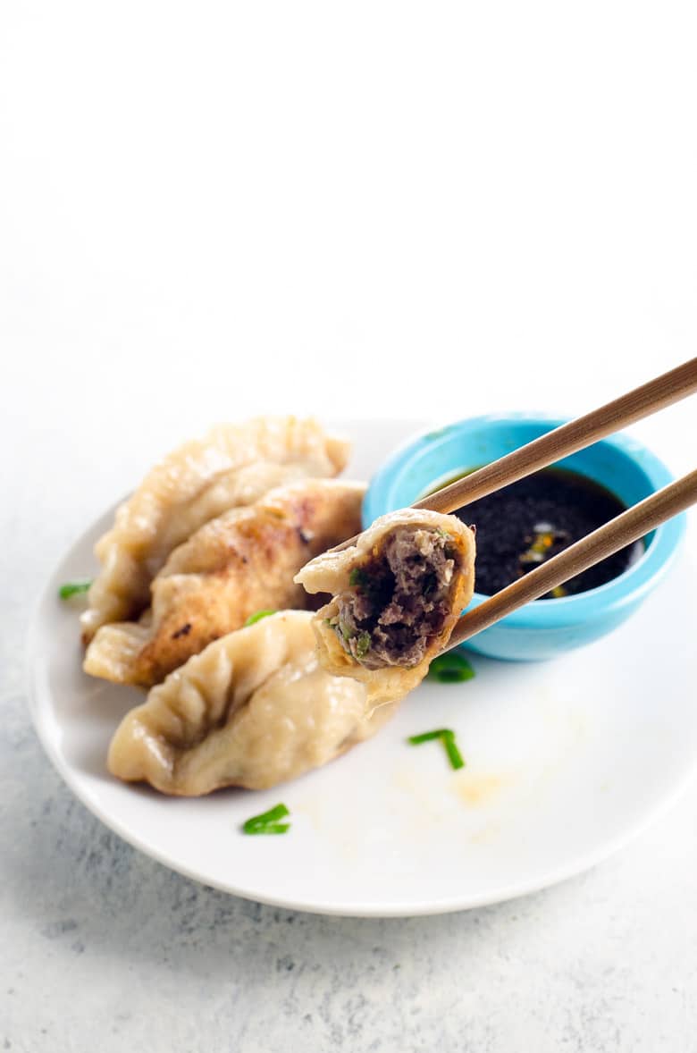 pork dumplings with a bite taken out of one of them, being held up by chopsticks over a white plate and a blue mise en place bowl with dumpling sauce