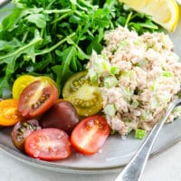 Tuna salad, rainbow tomatoes, arugula, and a lemon wedge on a grey plate on a light colored background