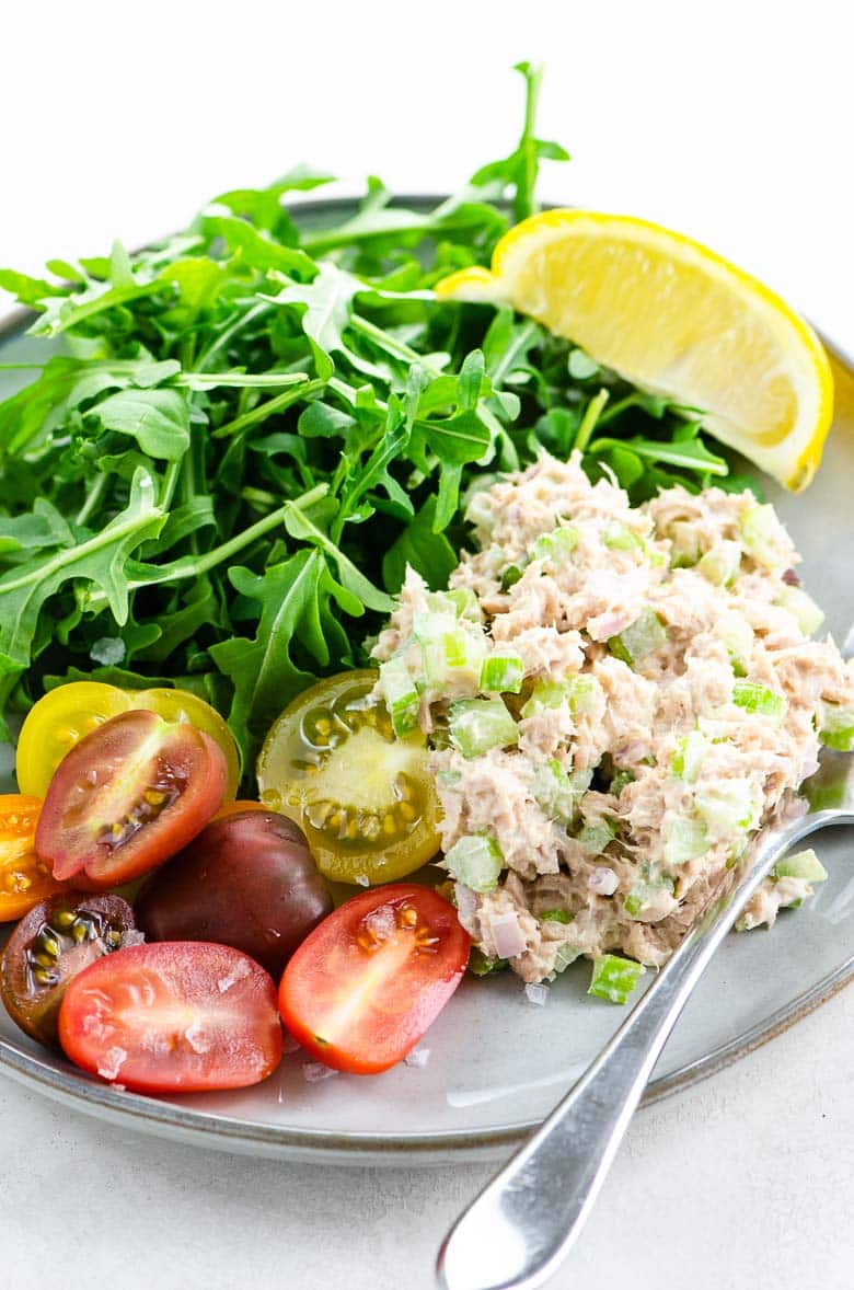 Tuna salad with capers, rainbow tomatoes, arugula, and a lemon wedge on a grey plate on a light colored background