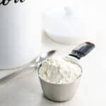 a cup of flour waiting to be leveled, a spoon, and a ceramic flour container with a lid