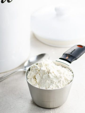 Heaping cup of flour with spoon and jar of flour in background