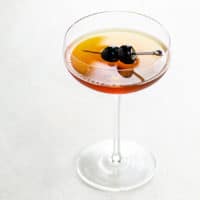 Coupe glass on a white background with a golden brown cocktail and two skewered luxardo cherries
