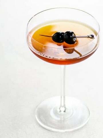 Coupe glass on a white background with a golden brown cocktail and two skewered luxardo cherries