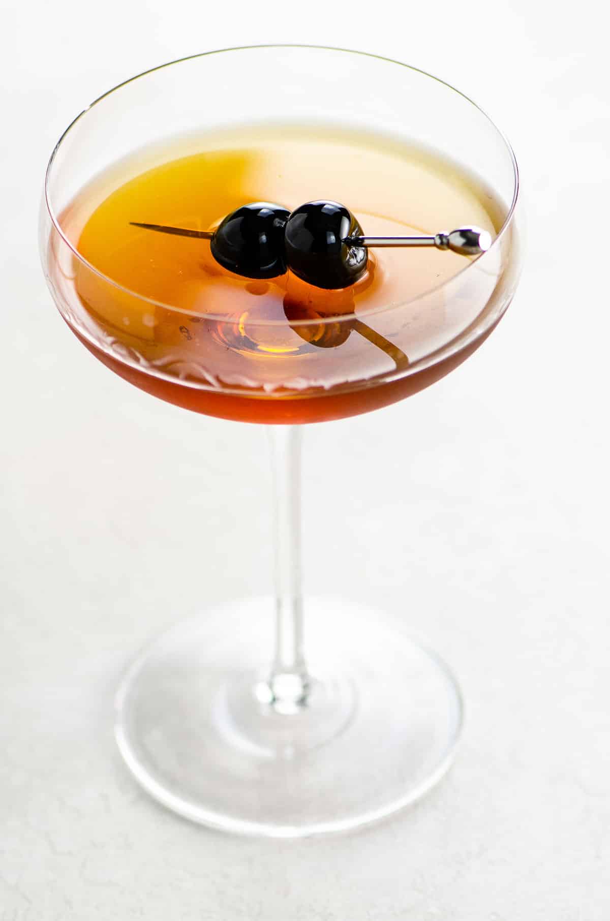 a manhattan cocktail in a coupe glass garnished with two speared luxardo cherries