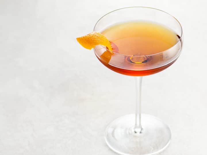 brown cocktail in a coupe glass garnished with an orange twist on a light background