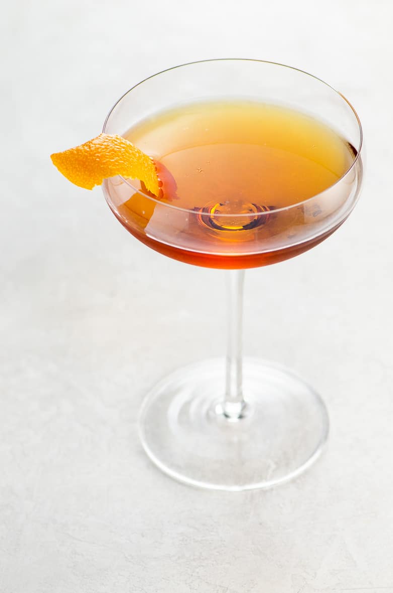brown cocktail in a coupe glass garnished with an orange twist on a light background