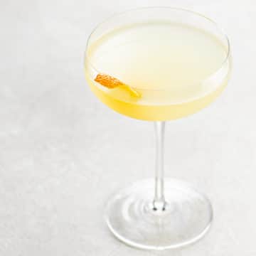 light orange tinted cocktail in a couple glass, garnished with an orange twist, on a light background