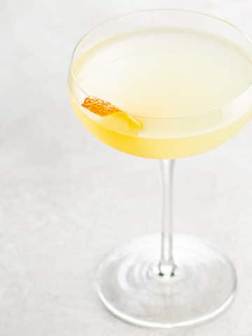 light orange tinted cocktail in a couple glass, garnished with an orange twist, on a light background