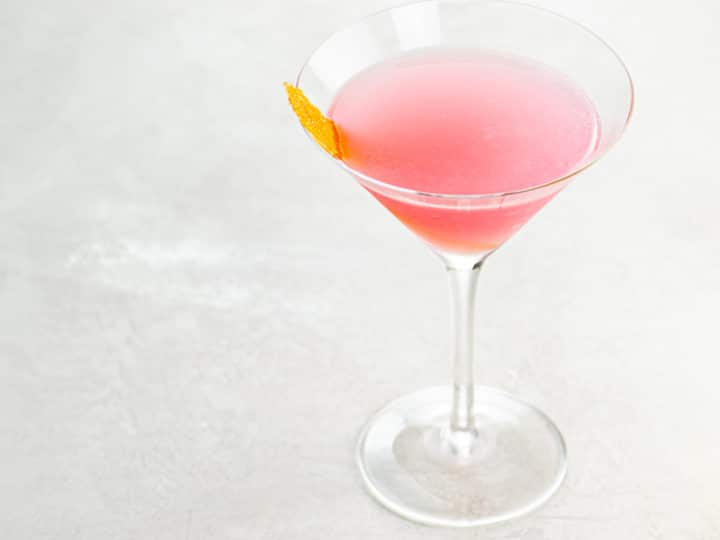 A pink cosmopolitan cocktail garnished with an orange twist in a cocktail glass on a light background