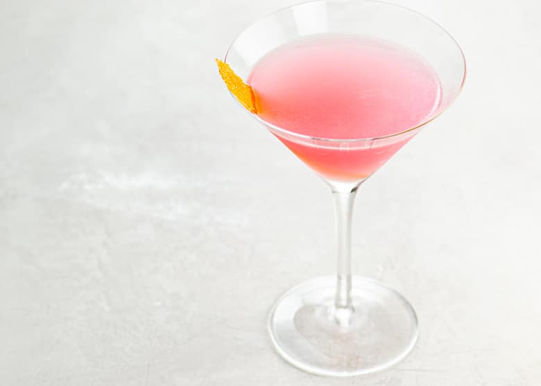 A pink cosmopolitan cocktail garnished with an orange twist in a cocktail glass on a light background
