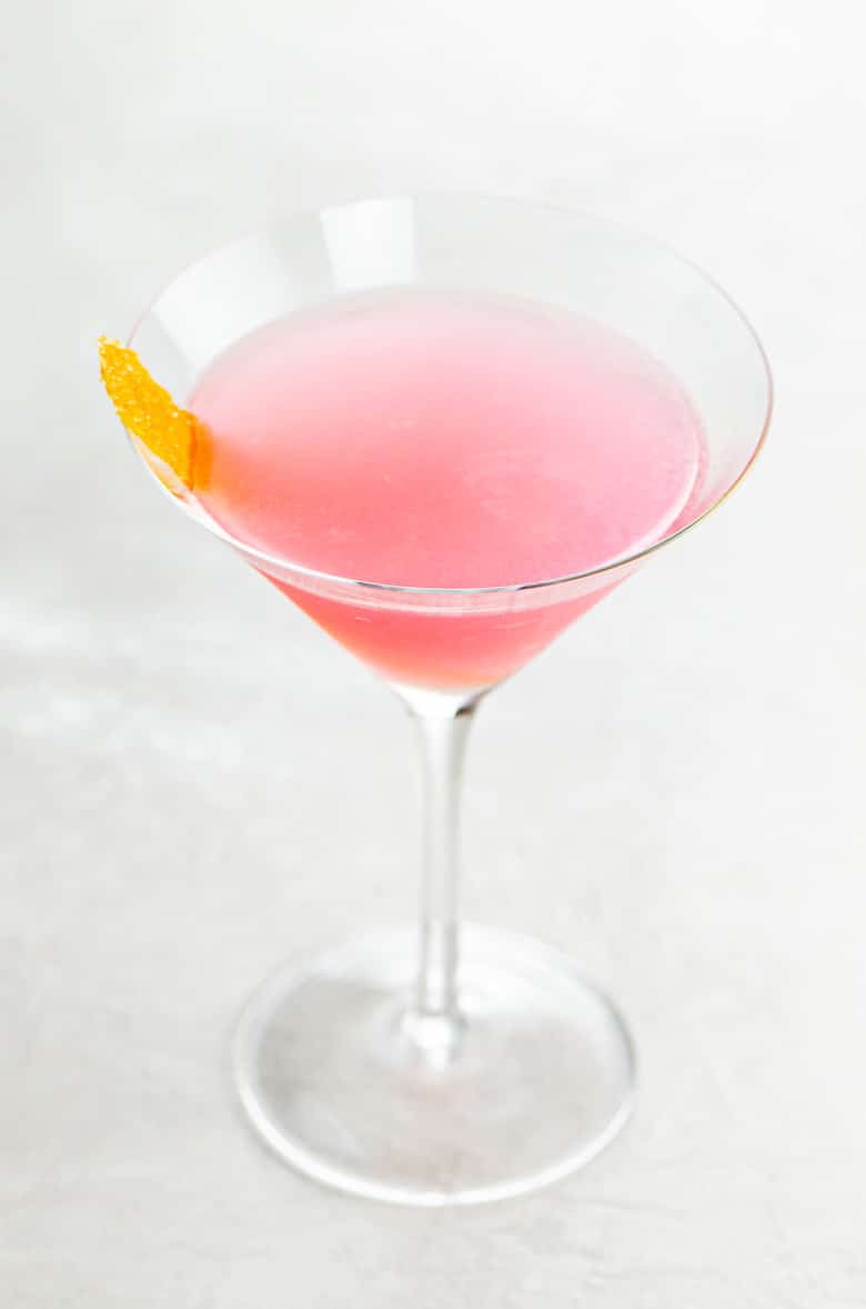 A pink cosmo cocktail garnished with an orange twist in a cocktail glass on a light background