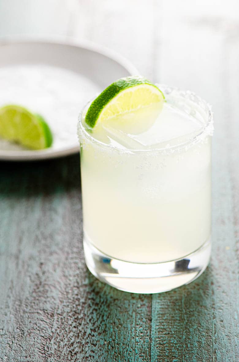 a margarita on the rocks glass garnished with a slice of lime on a blue wood background, with a plate and a lime wedge blurred in the background