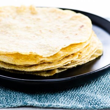 a stack of soft corn tortillas on a black plate on a blue textile background