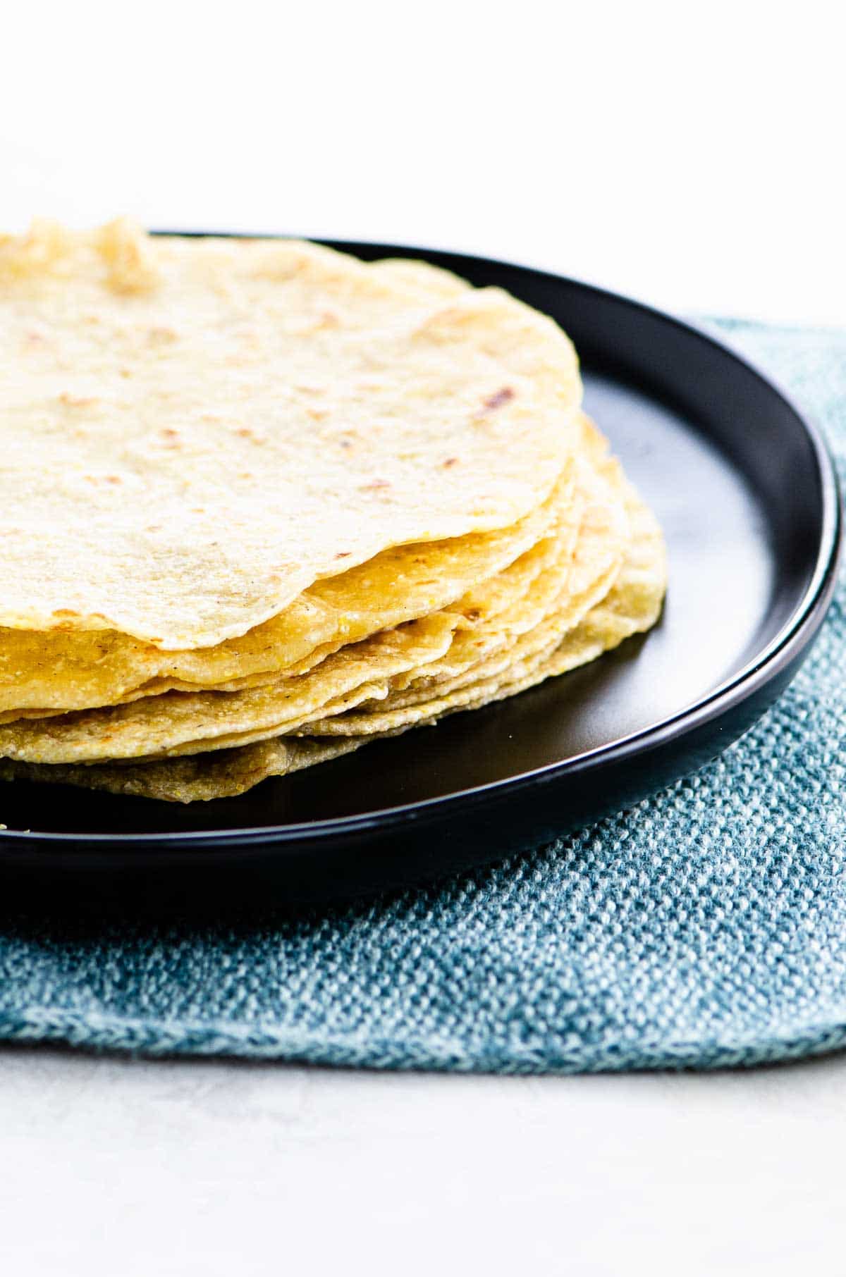 a stack of soft corn tortillas on a black plate on a blue textile background
