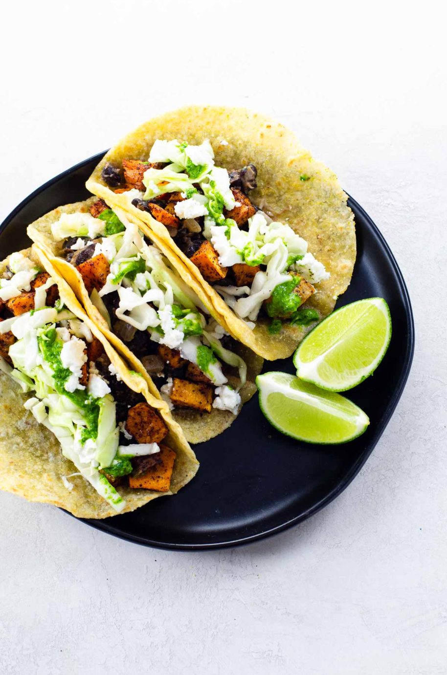 three tacos made with soft corn tortillas with two lime wedges on a black plate