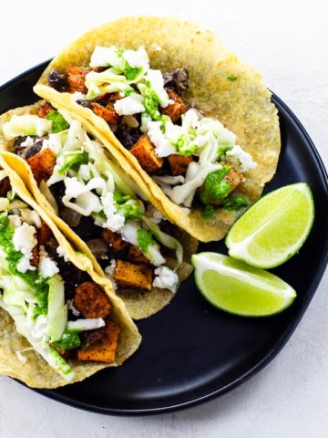 three tacos with two lime wedges on a black plate