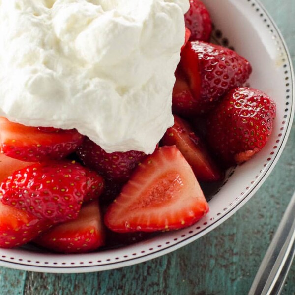 balsamic strawberries topped with whipped cream in a white bowl with a spoon on a blue background