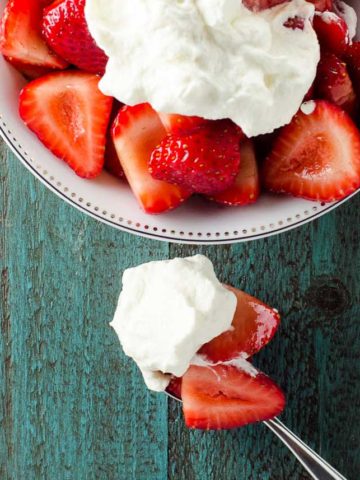balsamic strawberries topped with whipped cream in a white bowl with a spoon on a blue background