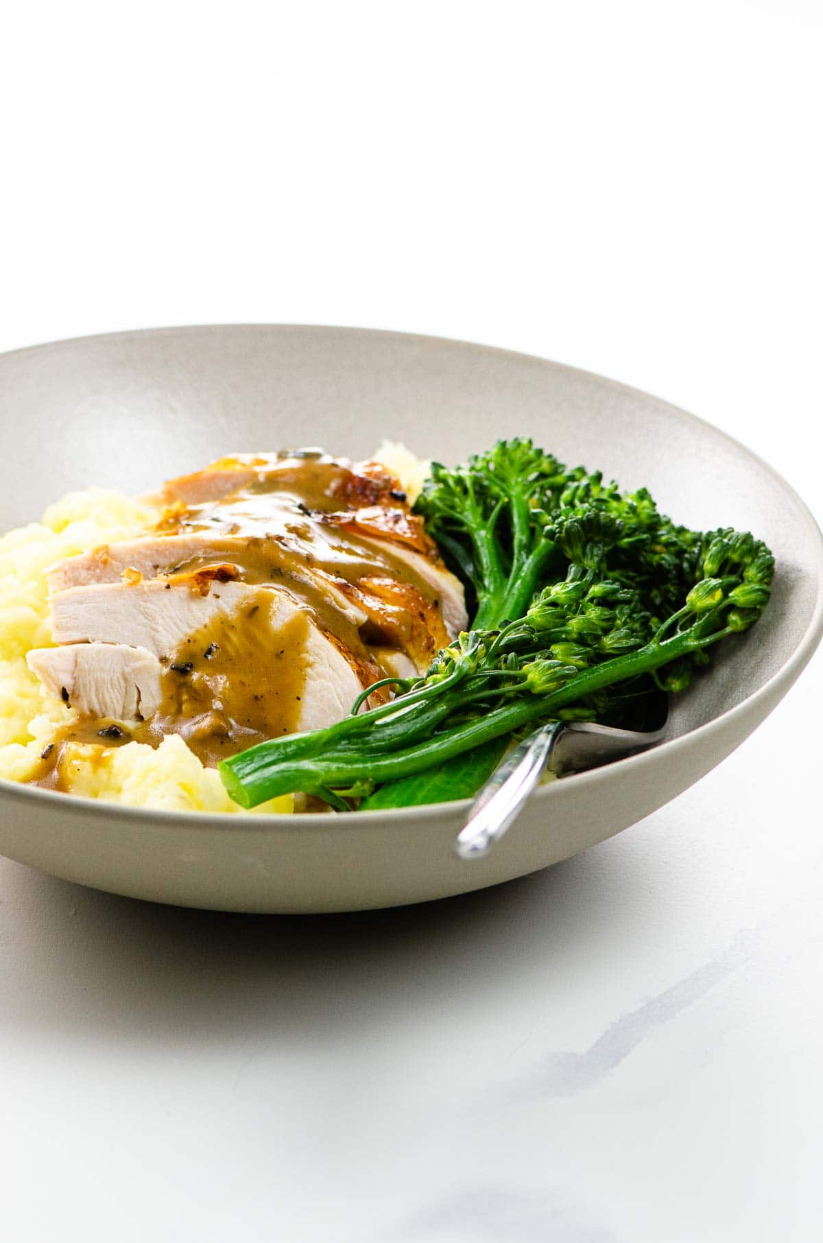 sliced old fashioned roast chicken with gravy over mashed potatoes with broccolini in a bowl