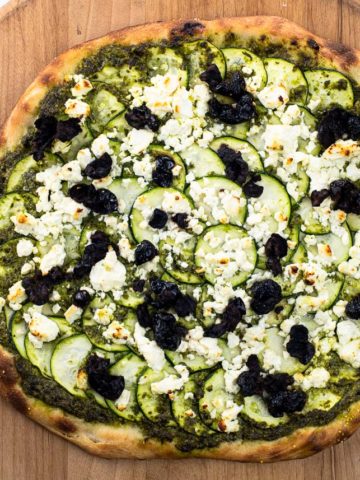pesto and feta pizza with zucchini and black olives on a pizza peel