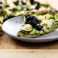 pesto and feta pizza with zucchini and black olives on a pizza peel and a plate