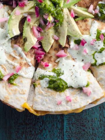 a refried bean quesadilla topped with avocado, sour cream, pickled onions, and sour cream on a blue background