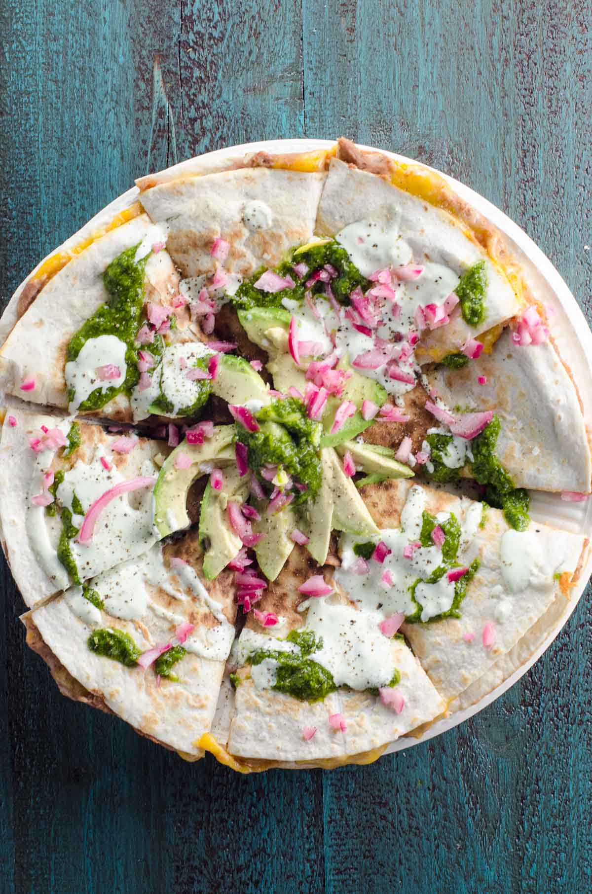 a refried bean quesadilla topped with avocado, sour cream, pickled onions, and sour cream on a blue background