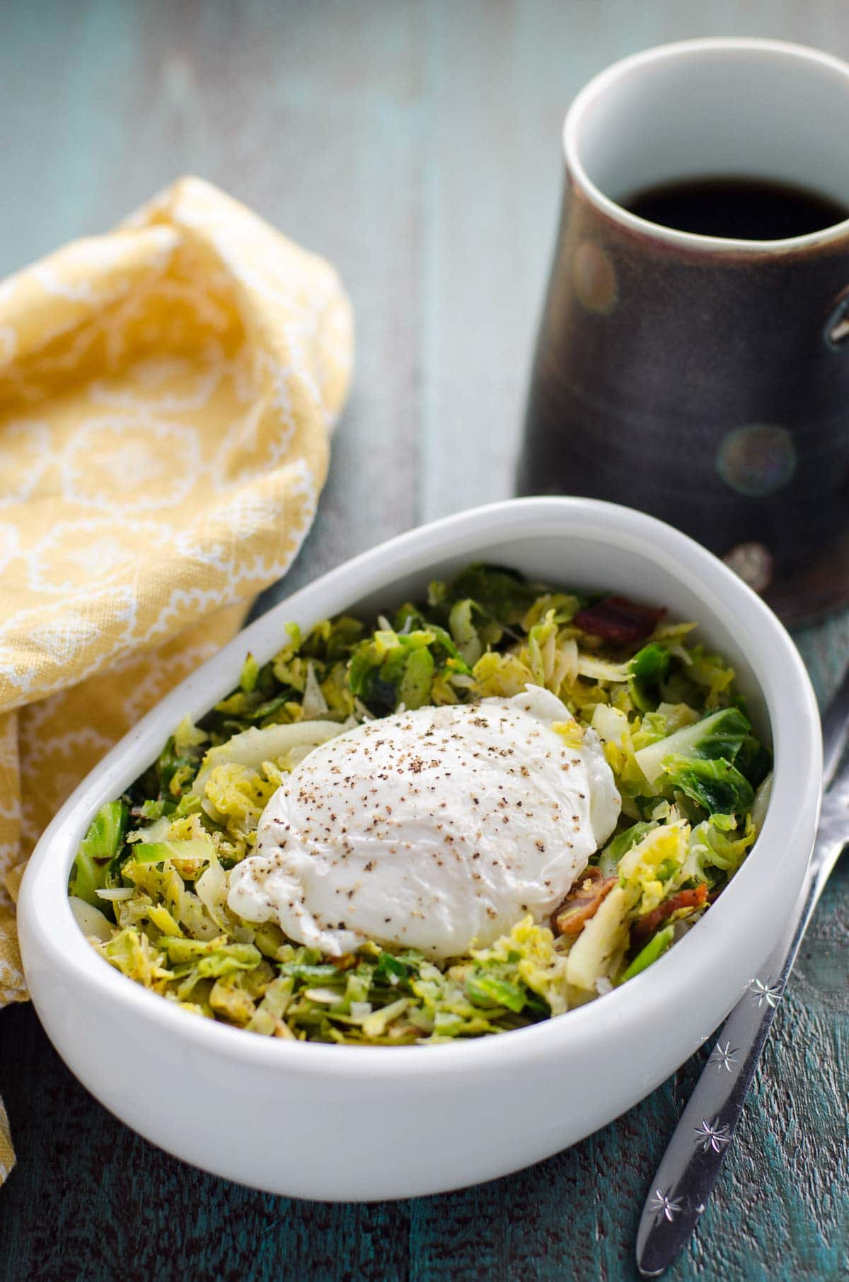 brussels sprouts hash with bacon and poached egg in a bowl on a table with coffee, a fork, and a napkin