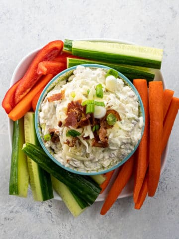 crack dip garnished with bacon, cheddar, and scallions in a serving bowl with carrots, cucumber, and red pepper sticks