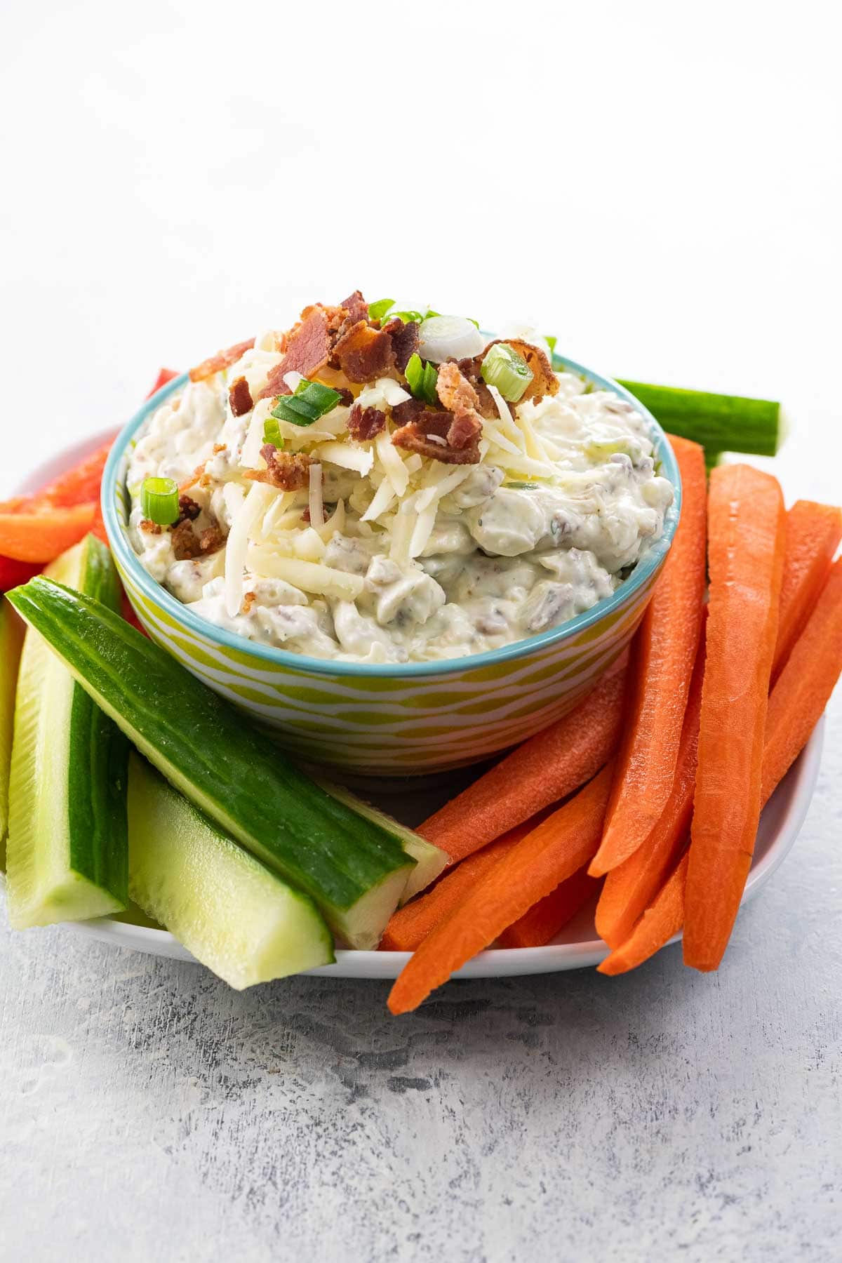 crack dip garnished with bacon, cheddar, and scallions in a serving bowl with carrots, cucumber, and red pepper sticks