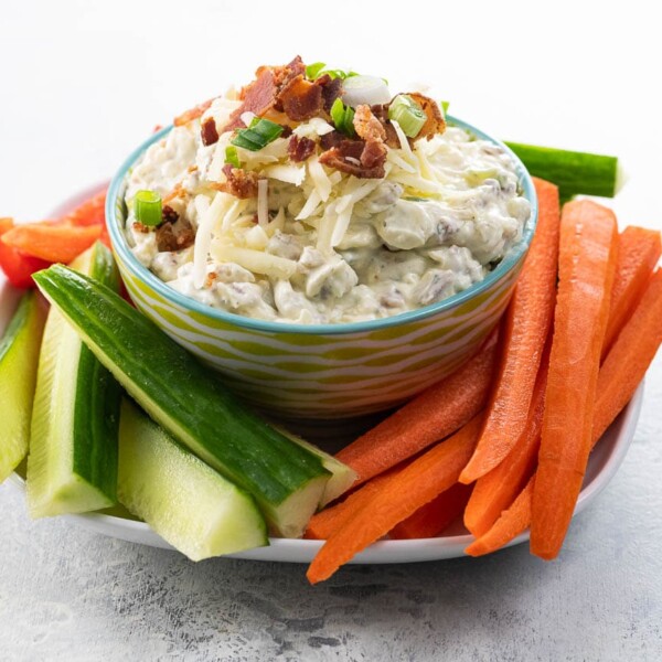 crack dip garnished with bacon, cheddar, and scallions in a serving bowl with carrots, cucumber, and red pepper sticks