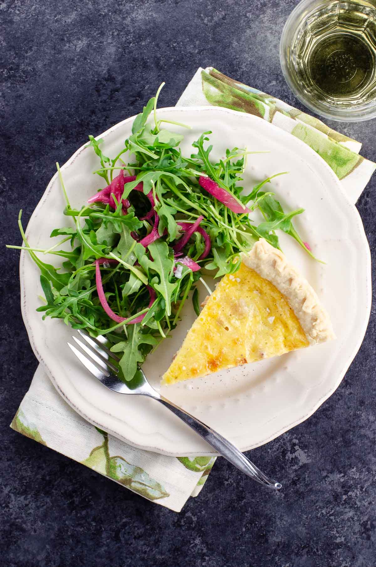 a slice of French Quiche Lorraine and salad on a plate, and a glass of white wine