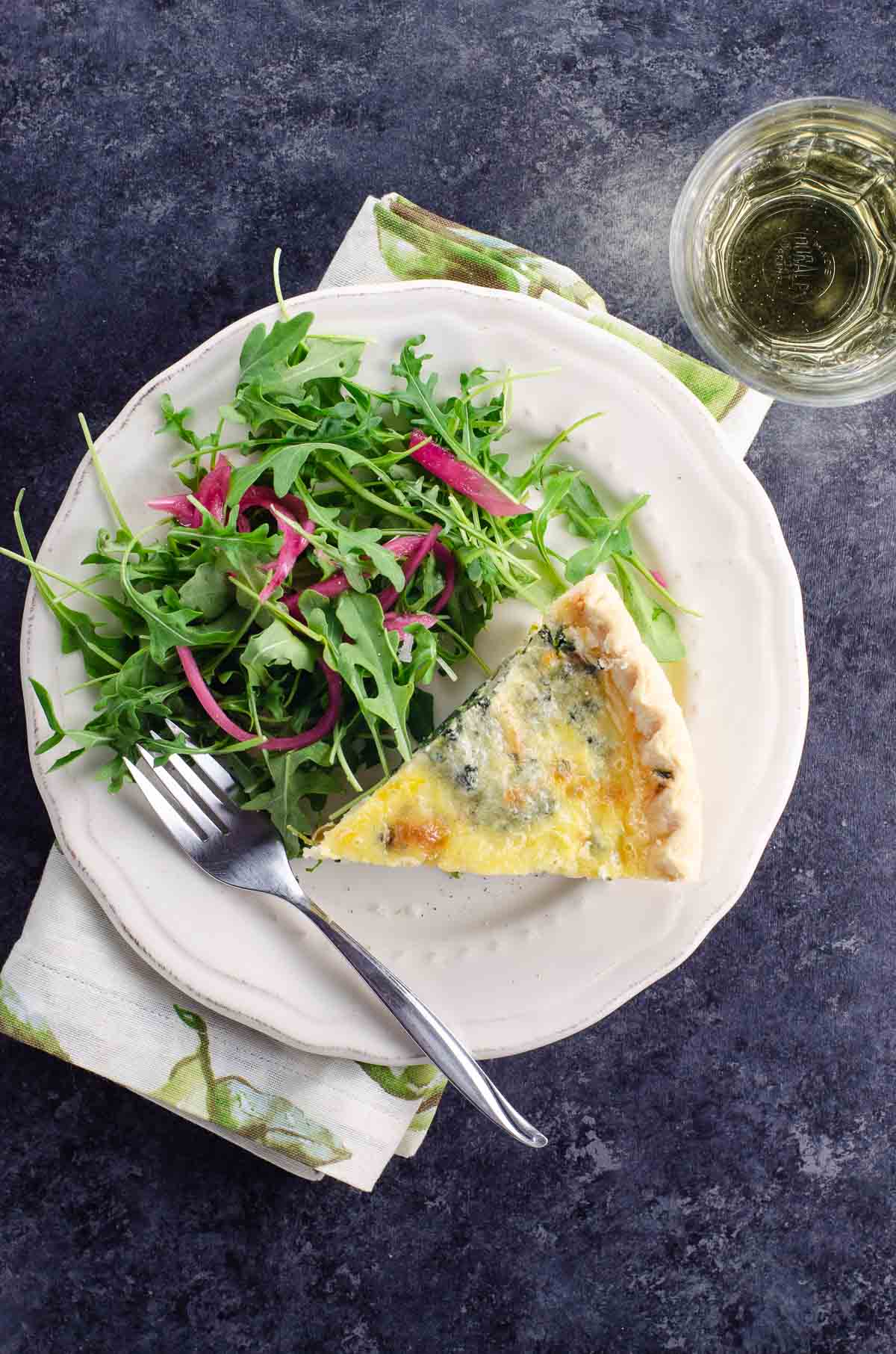 a slice of Quiche Florentine and salad on a plate, and a glass of white wine