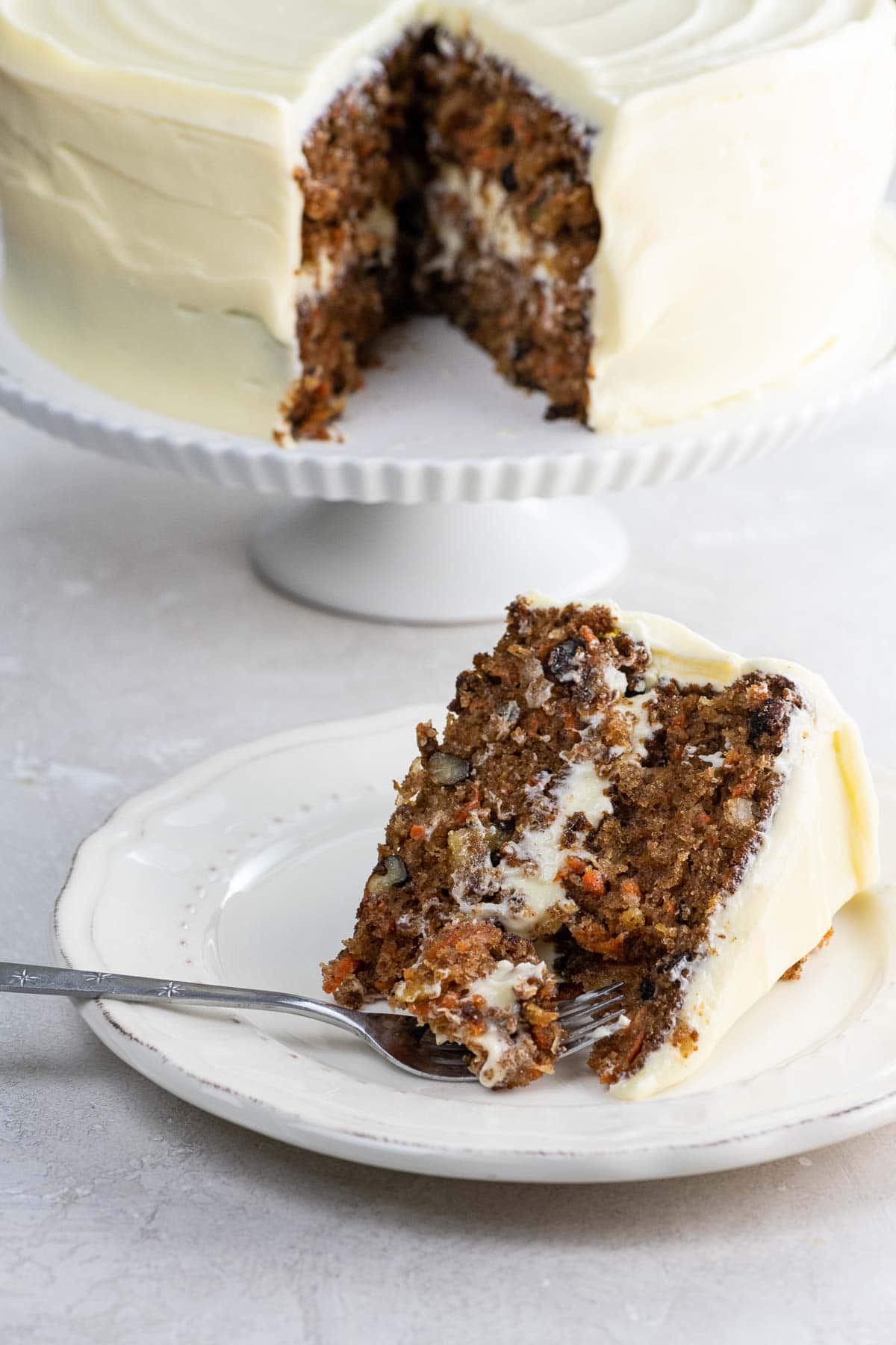 a slice of old fashioned carrot cake with not too sweet cream cheese frosting on a plate, with the whole cake behind it on a cake stand