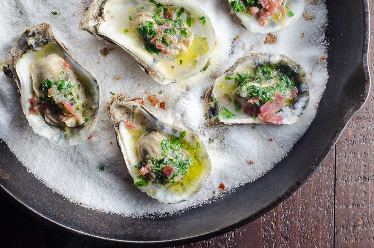 baked oysters with shallot herb butter and prosciutto on a bed of salt in a cast iron skillet