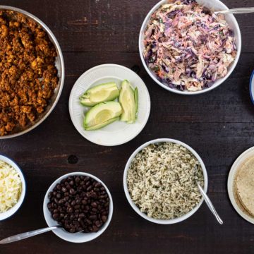 many ingredients for a taco bar laid out in bowls and on plates on a wooden surface