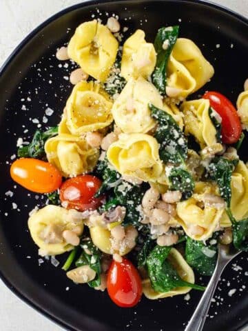 three cheese tortellini with white beans and vegetables on a black plate with a fork