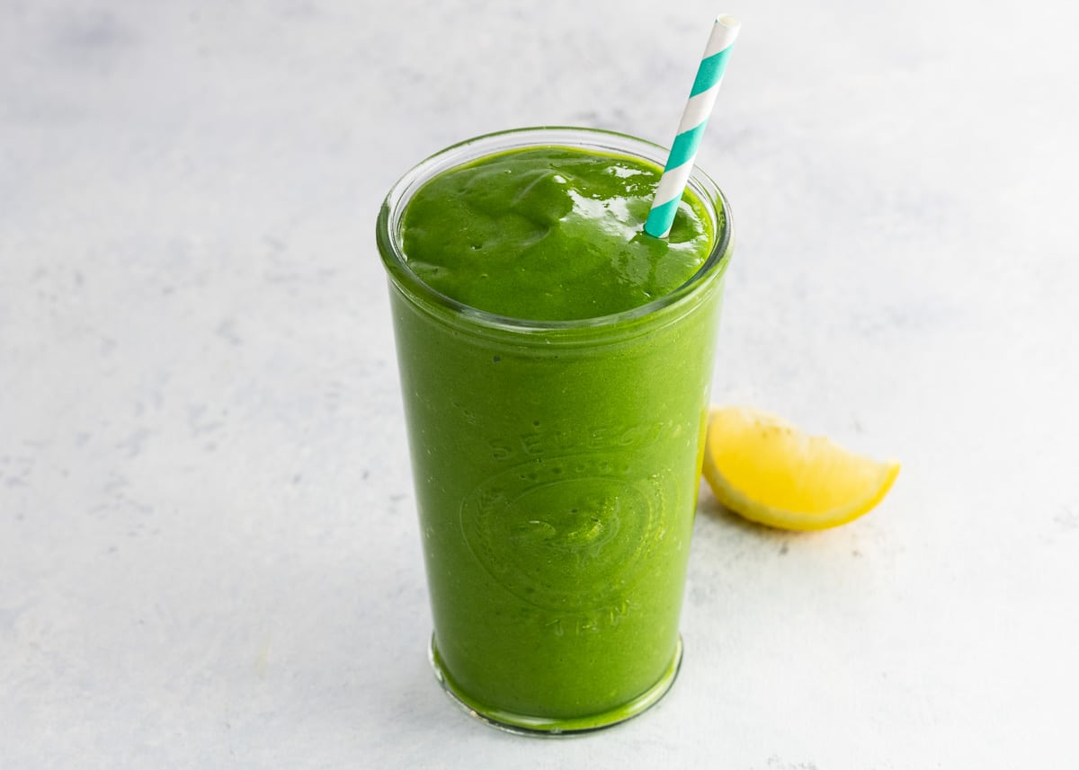 a mango and arugula smoothie in a glass with a paper straw and a lemon wedge