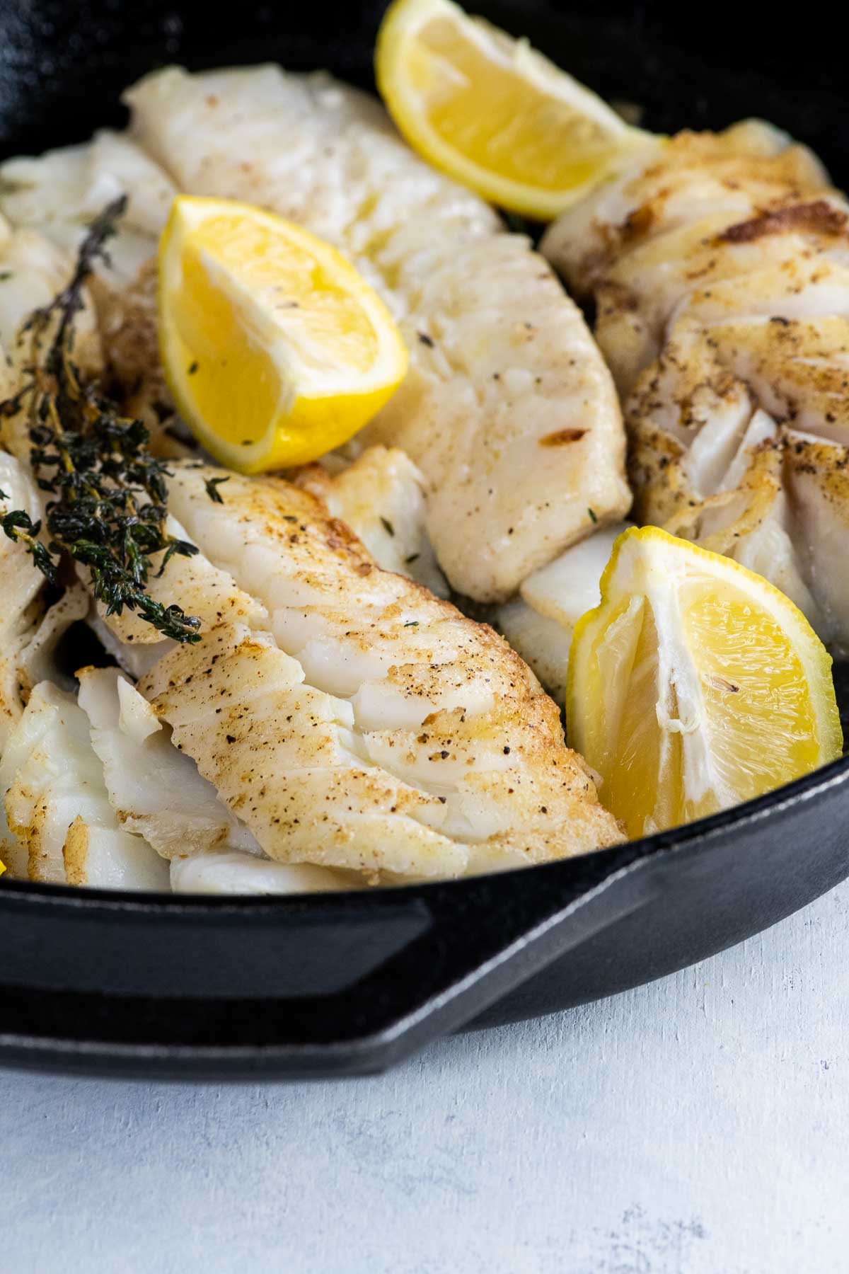 pan fried cod with thyme and lemon wedges in a cast iron skillet