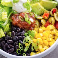 vegan taco bowl with black beans, corn, avocado, salsa, hot sauce, olives, lettuce, scallions, and vegan sour cream
