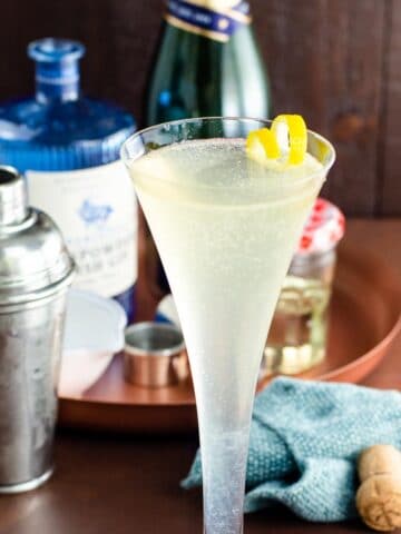 a soixante quinze gin champagne cocktail (french 75) in a glass with a lemon twist on a table with ingredients behind it