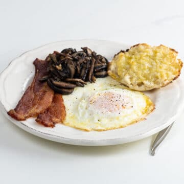 a fried egg over medium with bacon, sauteed mushrooms, and half an english muffin on a plate with a fork