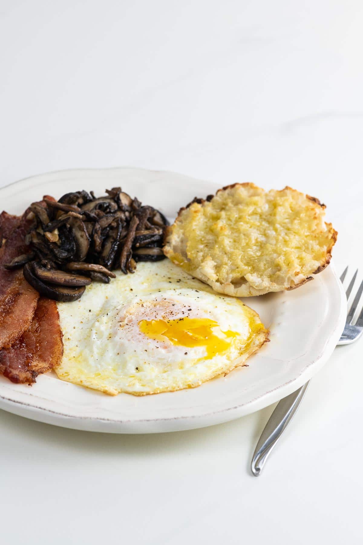 a fried egg over medium with bacon, sauteed mushrooms, and half an english muffin on a plate with a fork